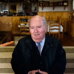 Retrato de Victoriano del Río Hijo, sentado en un sofá, con un fondo de madera y chimenea.