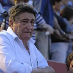Hombre mayor con camisa blanca, posible dueño de ganadería Domingo Hernández, observa atentamente en una plaza de toros.