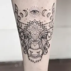 Un detallado tatuaje taurino con un ojo místico y fases lunares sobre la cabeza de un toro adornado con motivos florales y ornamentales.