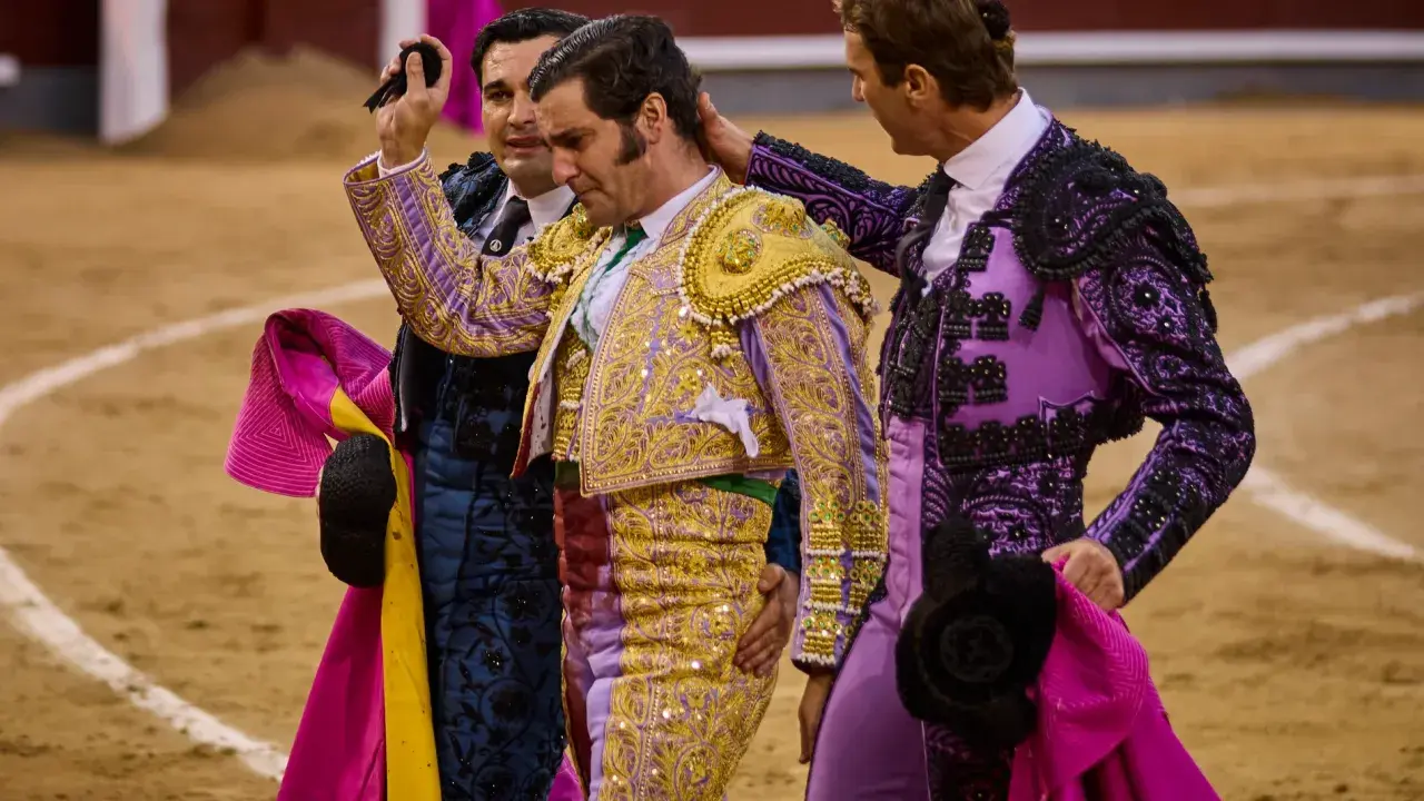 Tres toreros con trajes de luces, uno con coleta de torero, son felicitados en la arena.