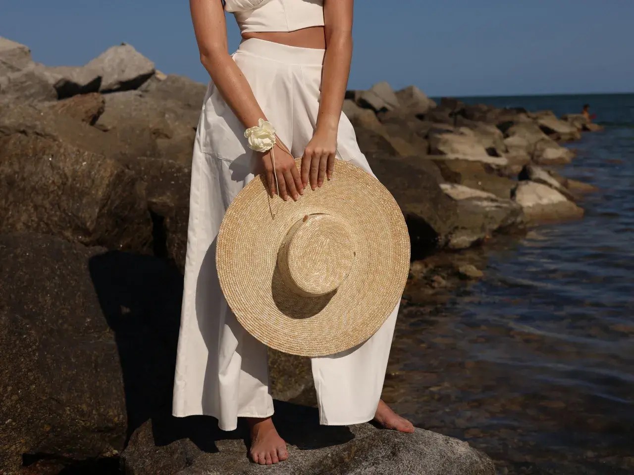 Mujer con un sombrero para ir a los toros, vestida de blanco, de pie sobre rocas junto al mar.