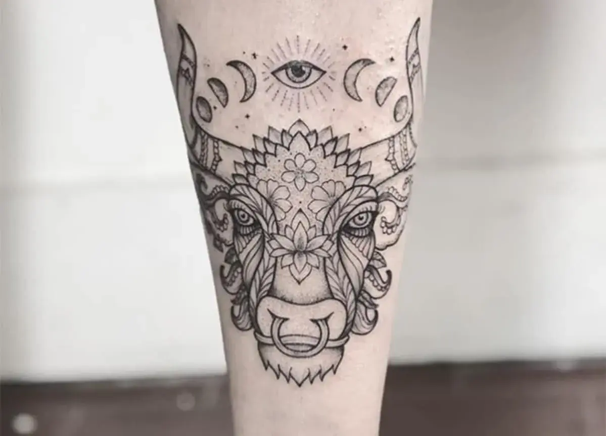 Un detallado tatuaje taurino con un ojo místico y fases lunares sobre la cabeza de un toro adornado con motivos florales y ornamentales.